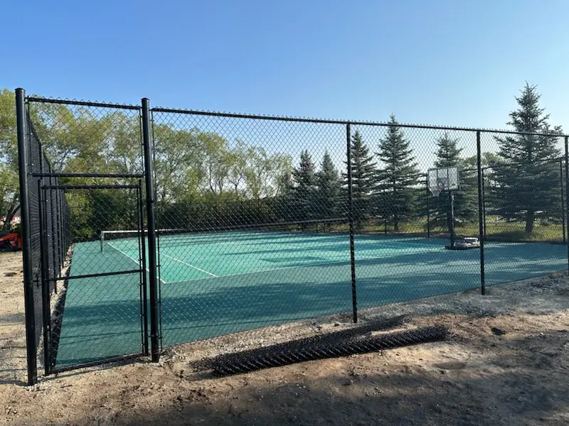 IMG_33771 (1) A fenced outdoor tennis court surrounded by trees under a clear sky.