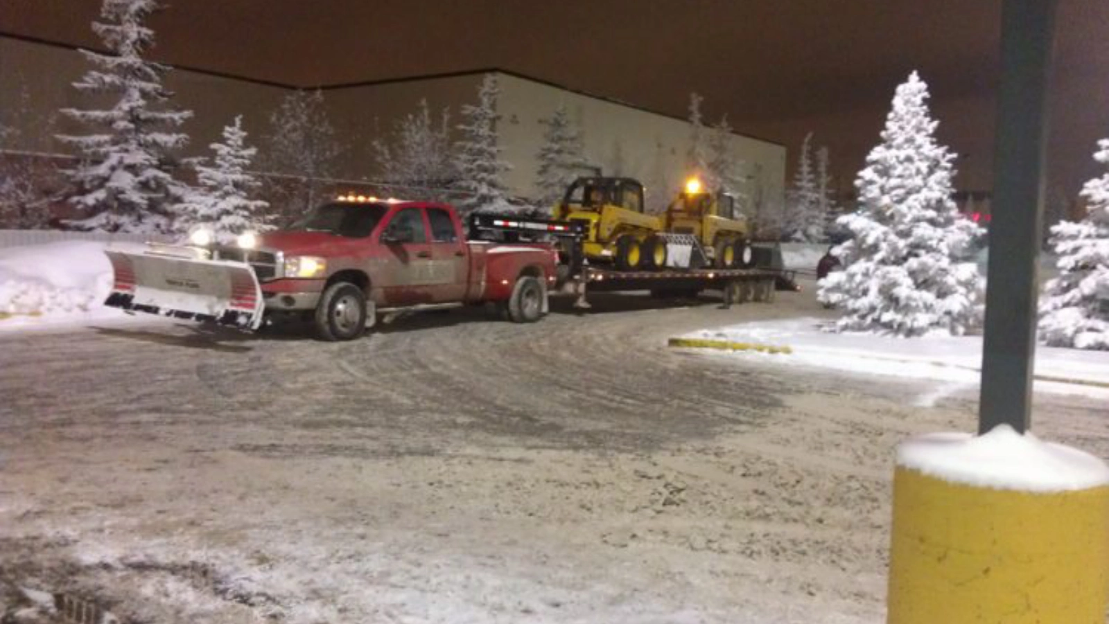 A red pickup truck towing a yellow excavator in a snowy nighttime setting.