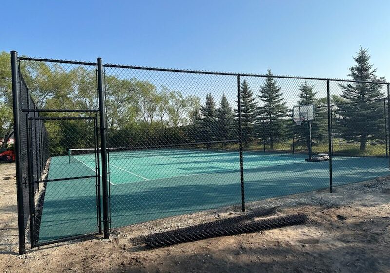 A fenced outdoor tennis court surrounded by trees under a clear sky.